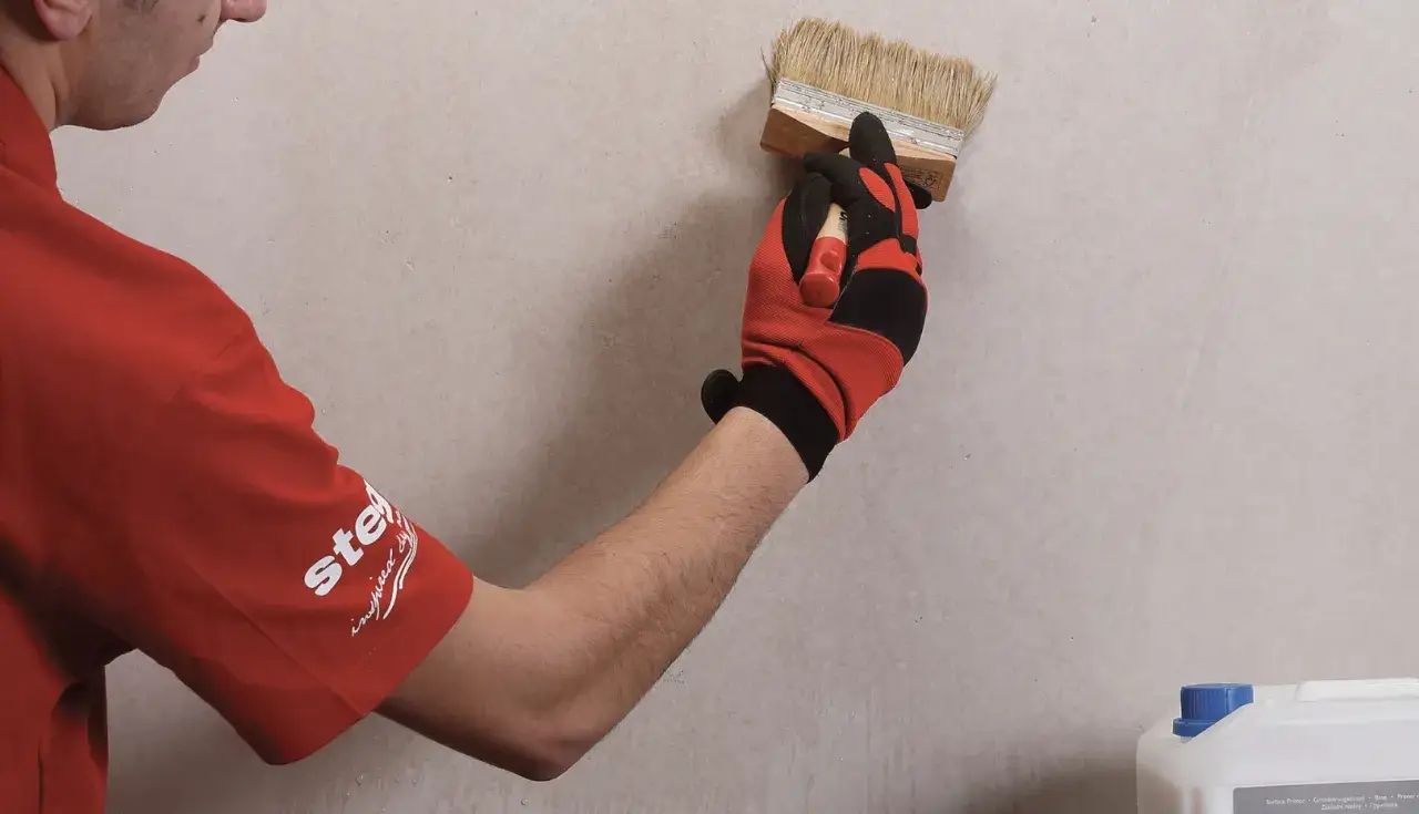 Gruntowanie płyt karton gips to kluczowy etap przygotowania powierzchni. Ręka w rękawicy nakłada preparat pędzlem na ścianę.