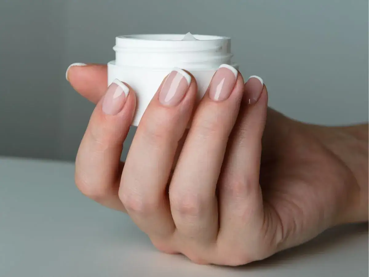 Dłoń z paznokciami w stylu francuskim trzyma słoiczek z kremem. Dowiedz się, jak nakładać bazę kauczukową dla idealnego manicure.