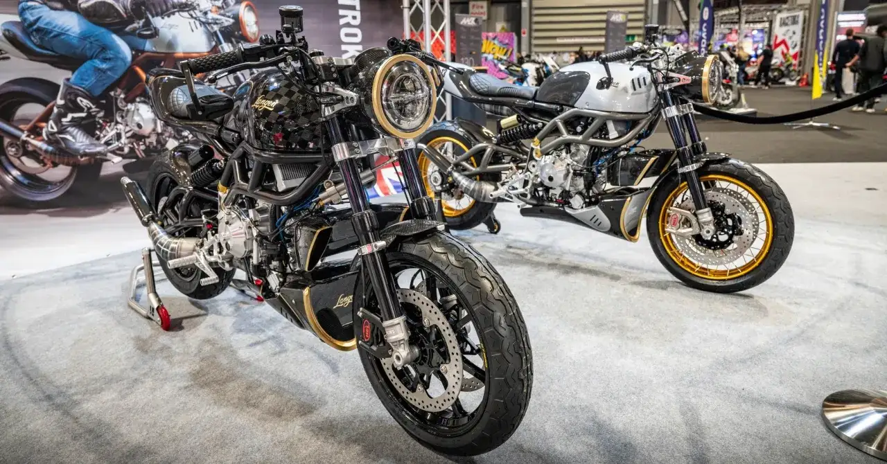 Dwa stylowe motocykle dwusuwowe, jeden czarny z elementami złota, drugi srebrny, prezentowane na wystawie.