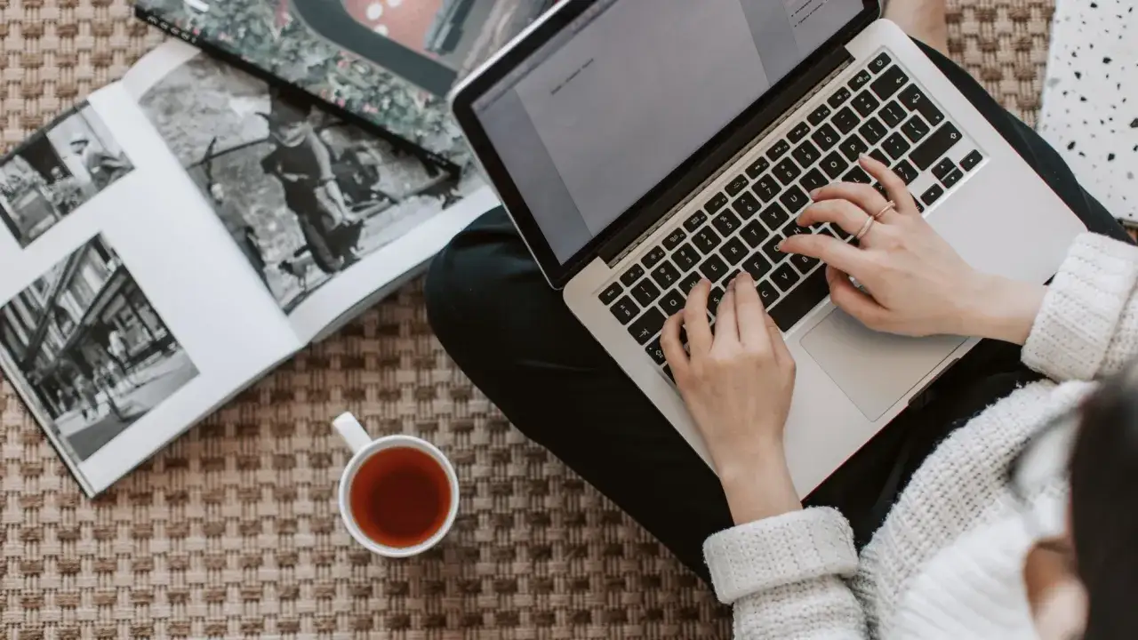 Kobieta pisze na laptopie, obok leży otwarta księga i filiżanka herbaty. Zastanawia się, ile kosztuje artykuł sponsorowany.