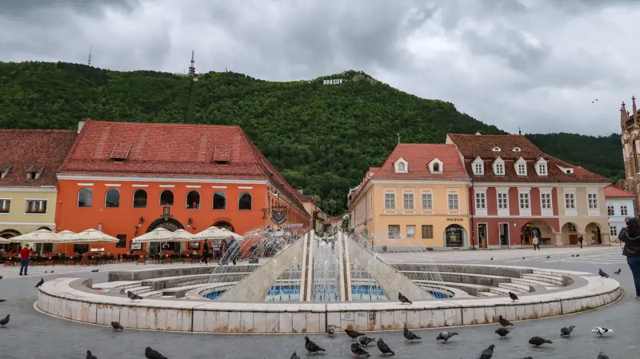 Fontanna na placu w Braszowie, z kolorowymi kamienicami i napisem "BRASOV" na wzgórzu. Wiele gołębi spaceruje wokół.