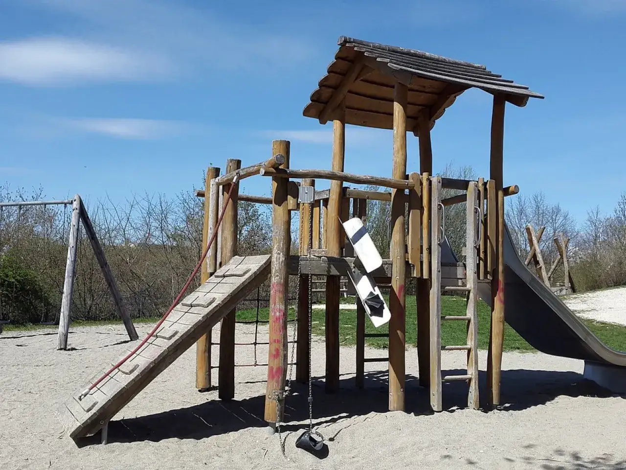 Drewniany plac zabaw ze zjeżdżalnią, drabinką i huśtawką na piaszczystym podłożu.