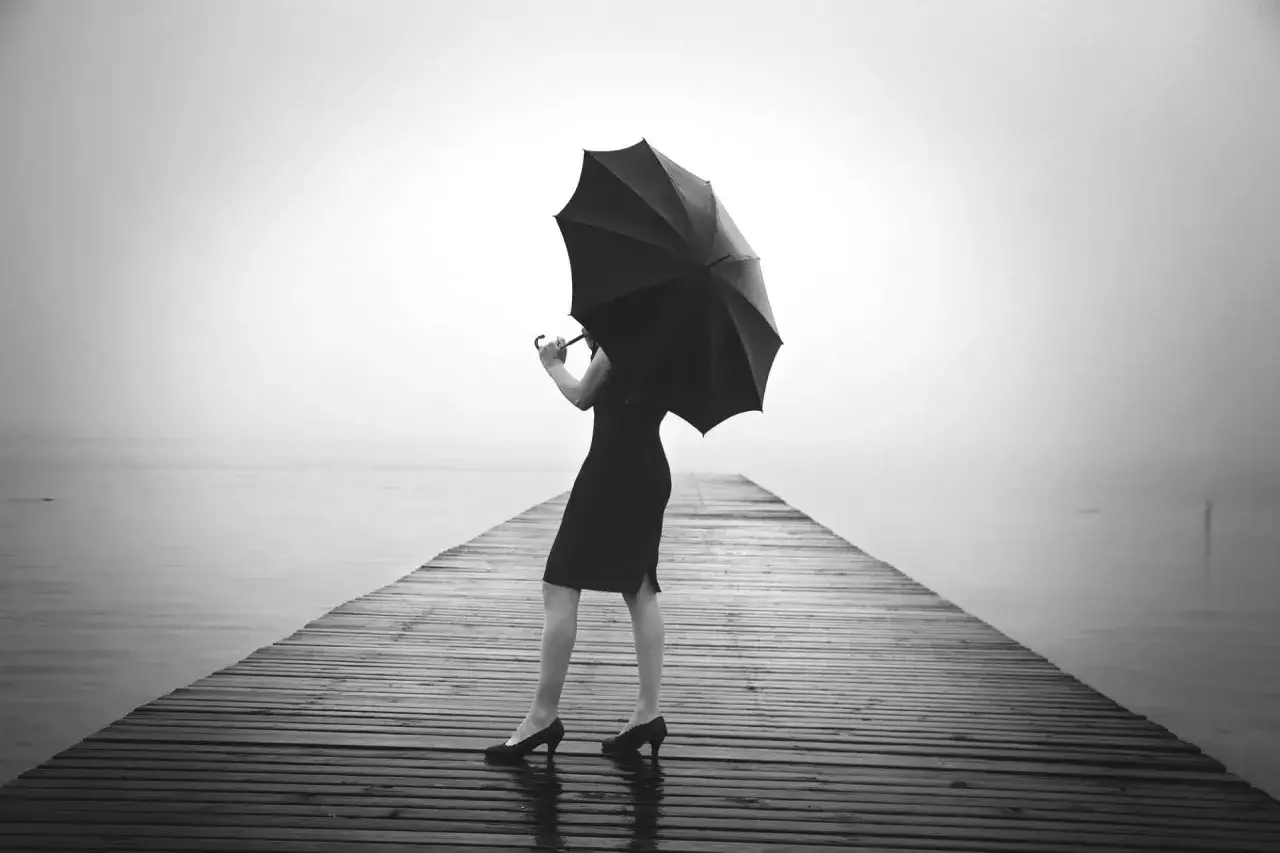 Kobieta w czarnej sukience stoi na drewnianym pomoście, trzymając parasolkę. W jej oczach widać myśli samobójcze, gdy patrzy w mglistą dal.