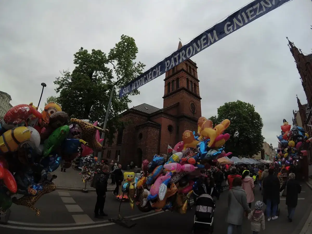 Kolorowe balony z postaciami z bajek i zwierzątkami, idealne gniezno atrakcje dla dzieci, na tle ceglanej wieży kościelnej i tłumu ludzi.