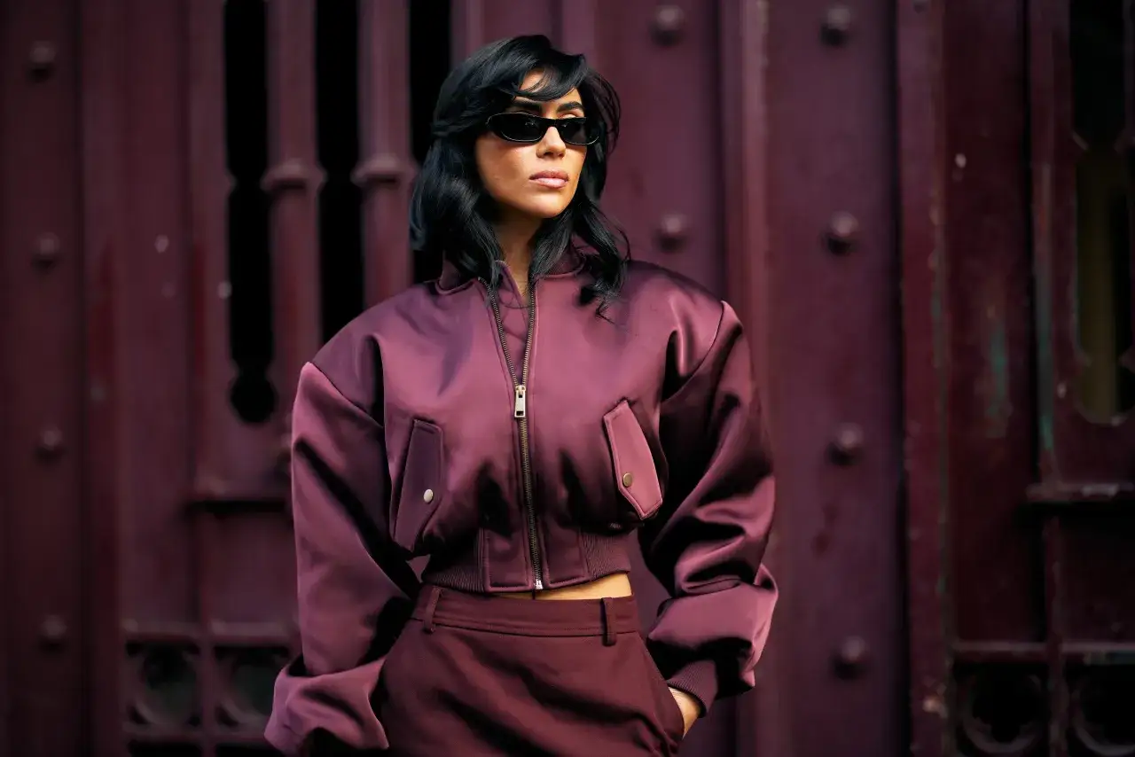 Kobieta w okularach przeciwsłonecznych i satynowej kurtce bomberce w odcieniu śliwki. Stylizacja nawiązuje do mody sprzed lat, z nutą retro.
