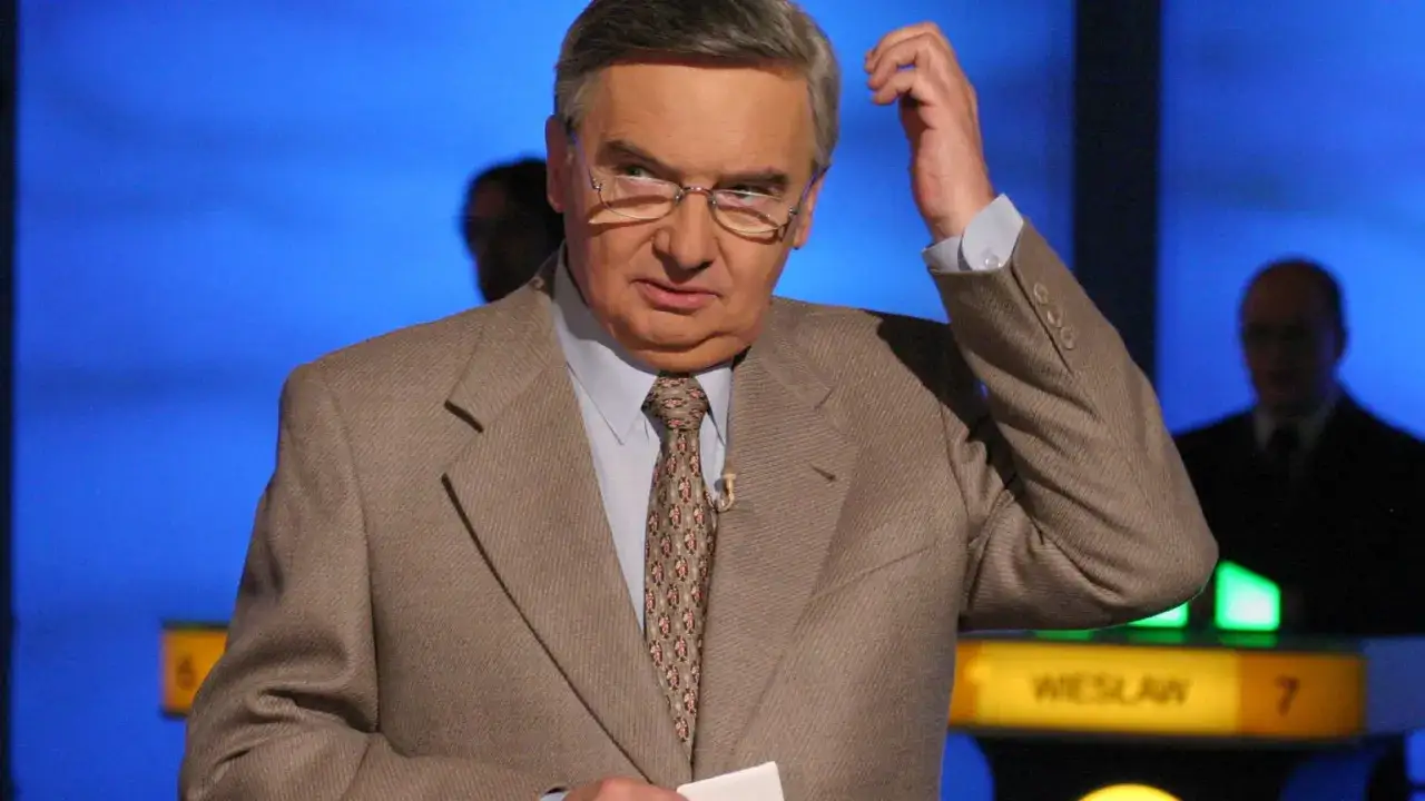 Tadeusz Szmuk, znany z teleturniejów, w garniturze i krawacie, z ręką przy głowie. Czy Tadeusz Szmuk żyje? Tak, jest aktywny.