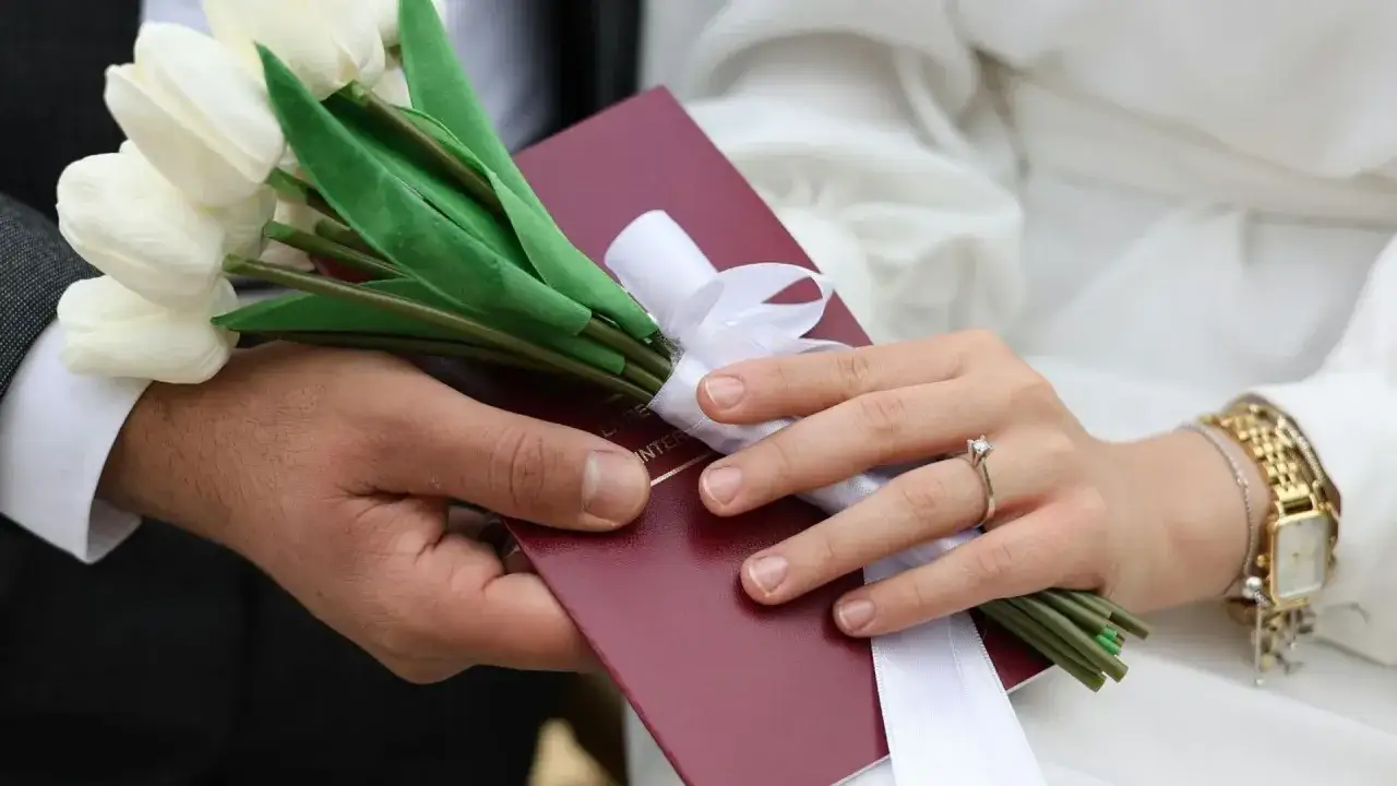 Dłoń mężczyzny i kobiety trzymają bukiet białych tulipanów i czerwony dokument.