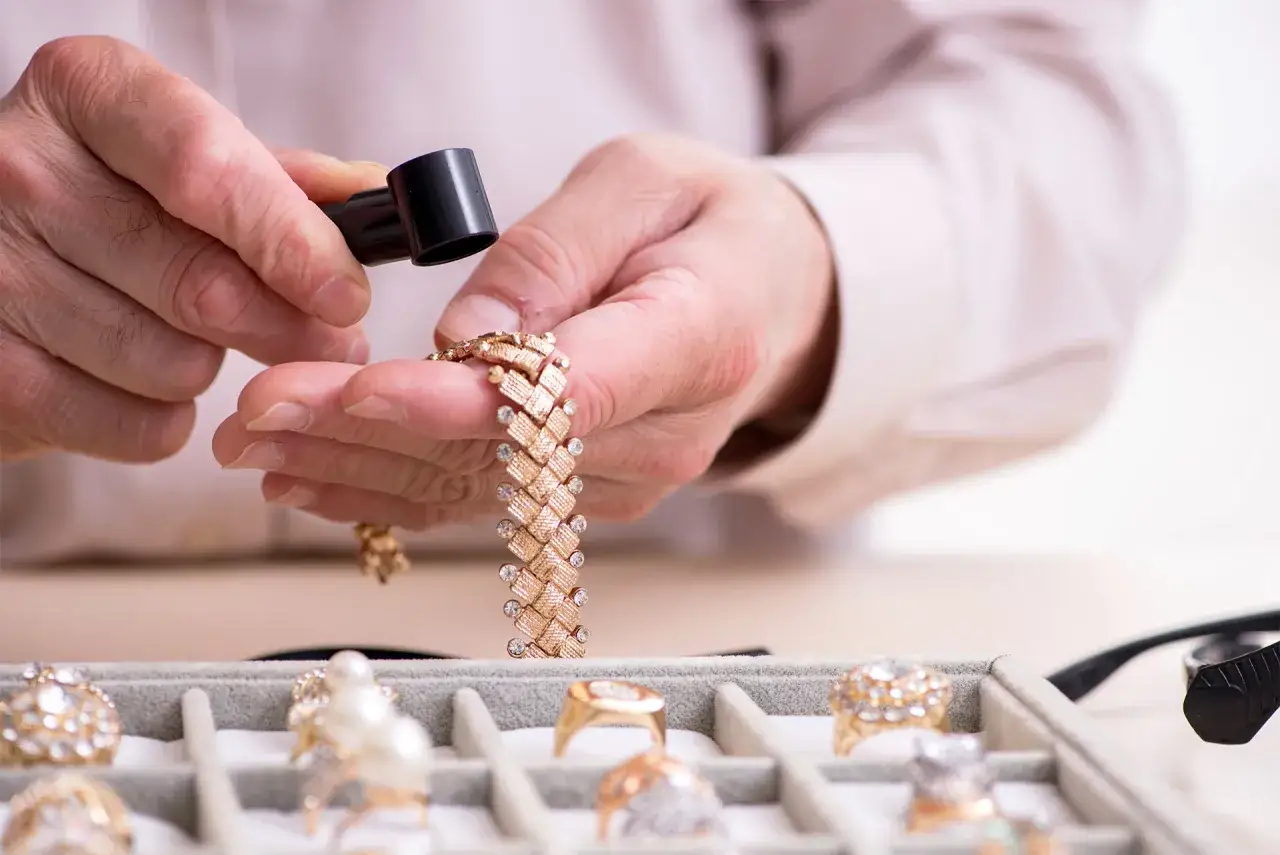 Dłonie mężczyzny trzymają złotą bransoletkę i lupę nad tacką z biżuterią.