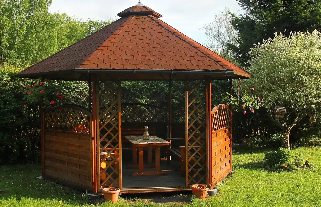Drewniana altana ogrodowa z czerwoną, łuskowatą papą na dachu, otoczona zielenią.
