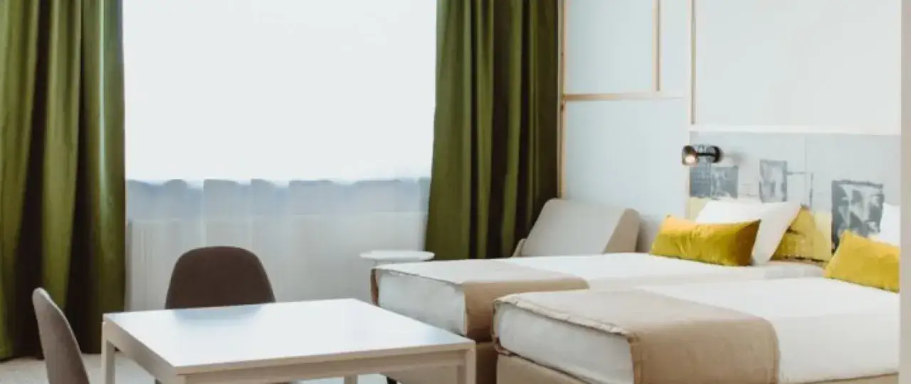 Pokój hotelowy z dwoma łóżkami, stołem i krzesłami, zielonymi zasłonami.