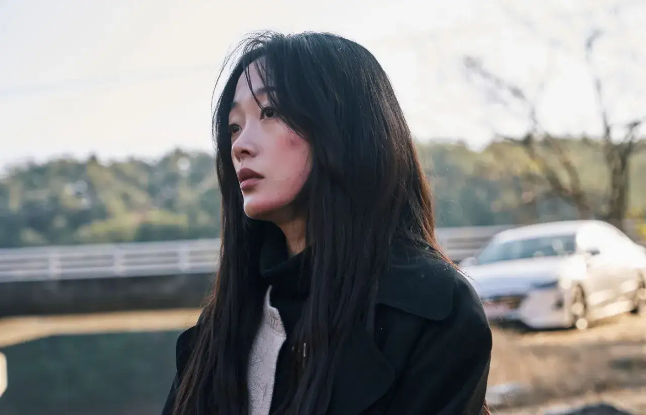 Młoda kobieta z zaczerwienionymi policzkami, wpatrzona w dal, gdy inni stali z boku. W tle widać samochód i most.