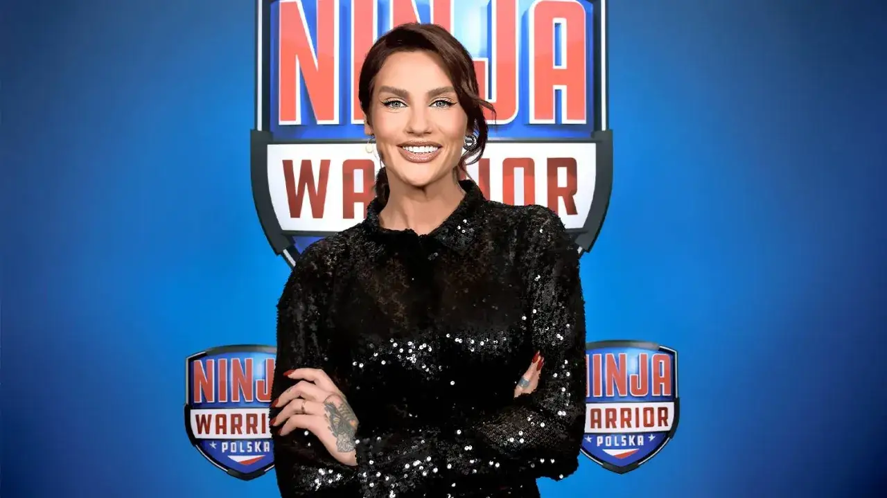 Uśmiechnięta prowadząca Ninja Warrior w błyszczącej, czarnej sukience, z tatuażami na rękach, na tle niebieskiego logo programu.