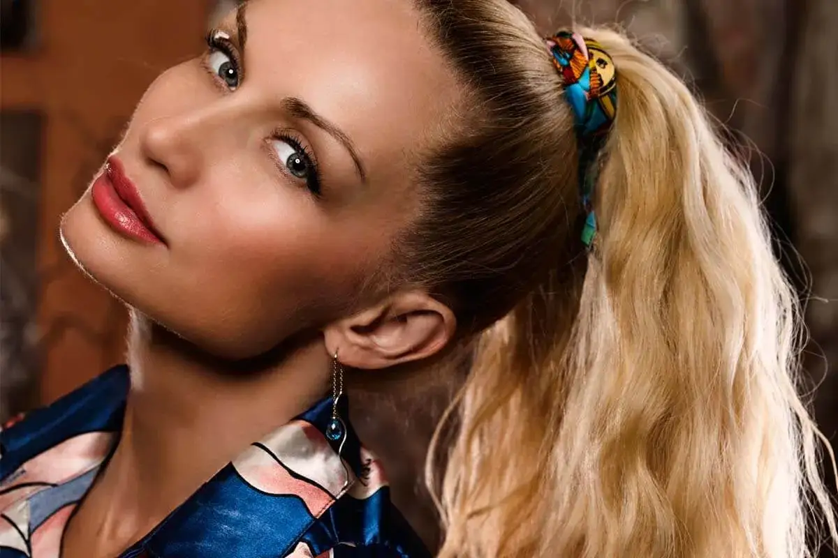 Kobieta z blond włosami spiętymi w kitkę z przedziałkiem, ozdobioną kolorową chustką.