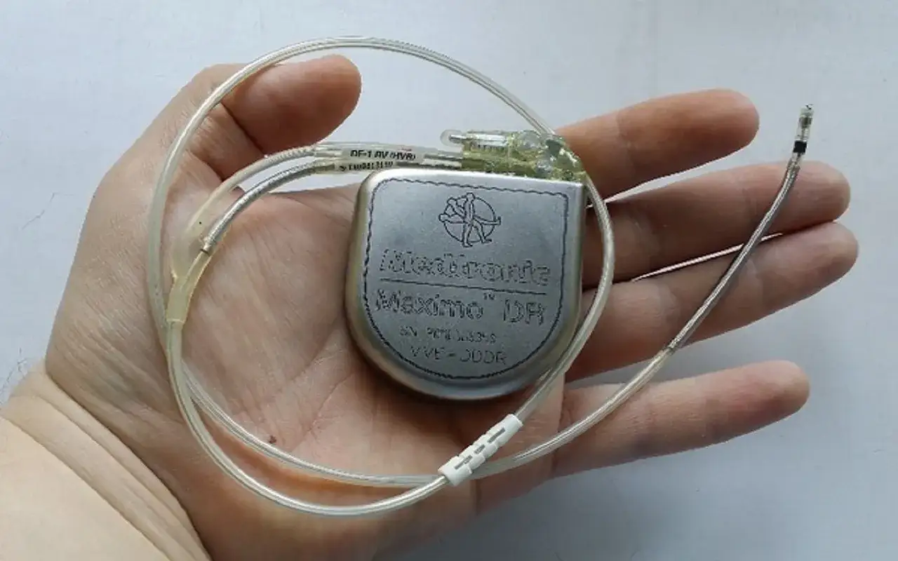 Dłoń trzyma kardiowerter Medtronic Maximo DR, urządzenie podobne do rozrusznika serca, z dwoma elektrodami.