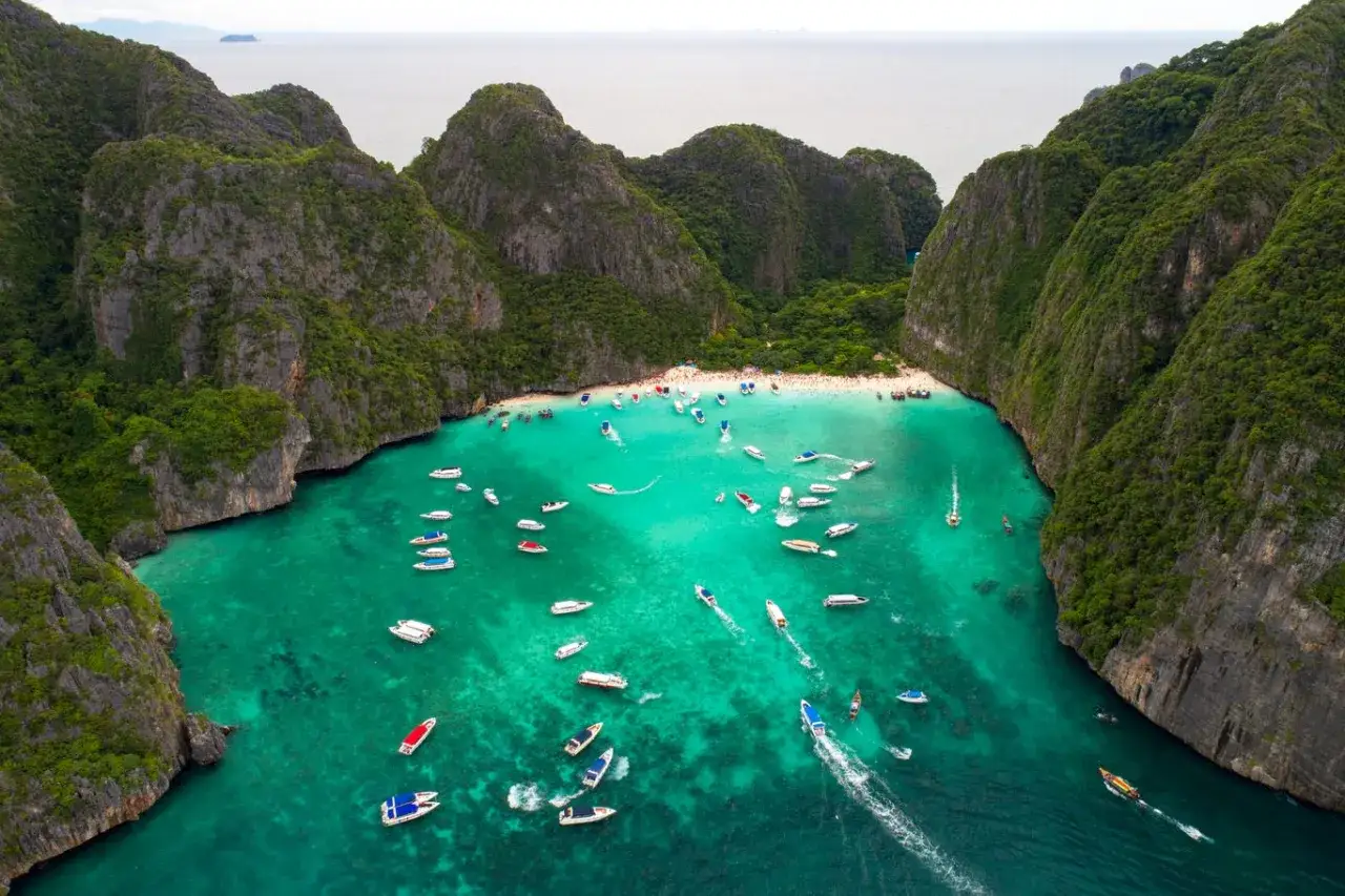 Maya Bay tętni życiem, pełna łodzi i turystów na piaszczystej plaży, otoczona stromymi, zielonymi klifami.