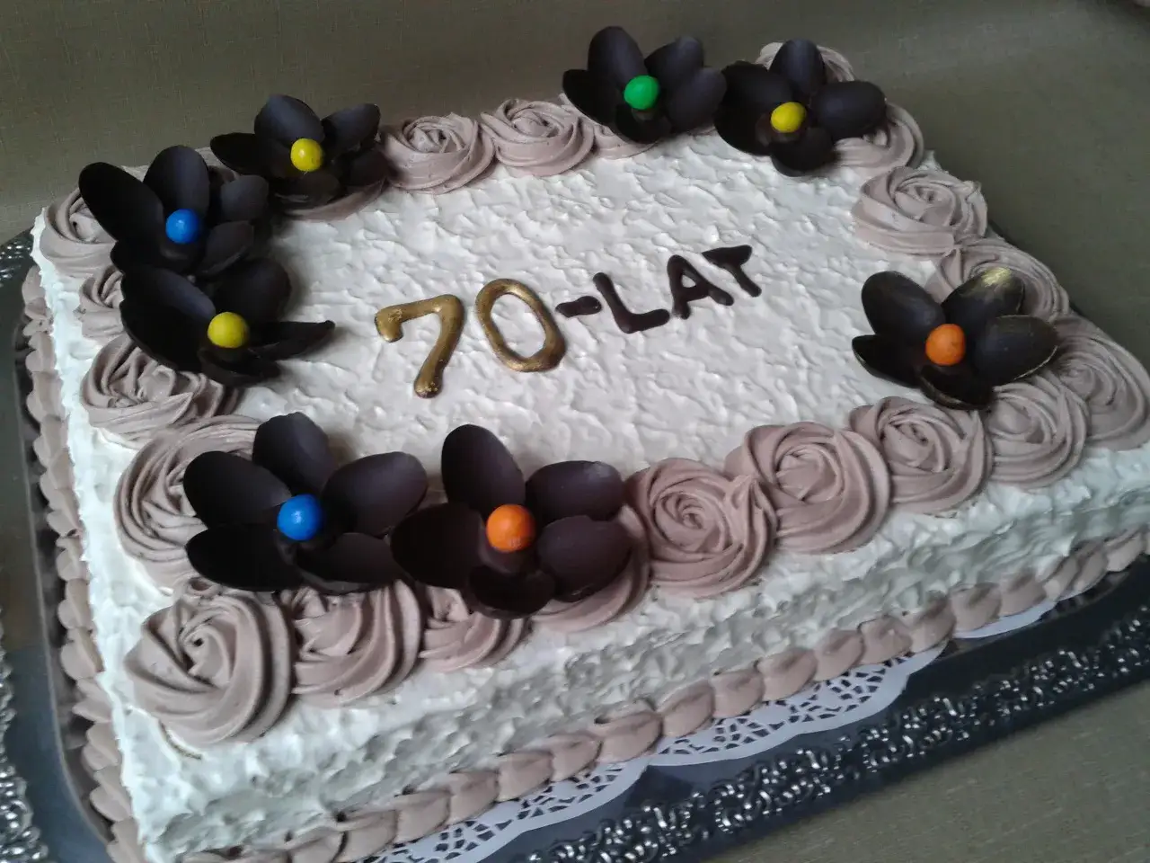 Prostokątny tort z białym kremem, ozdobiony czekoladowymi kwiatami i napisem "70-LAT".
