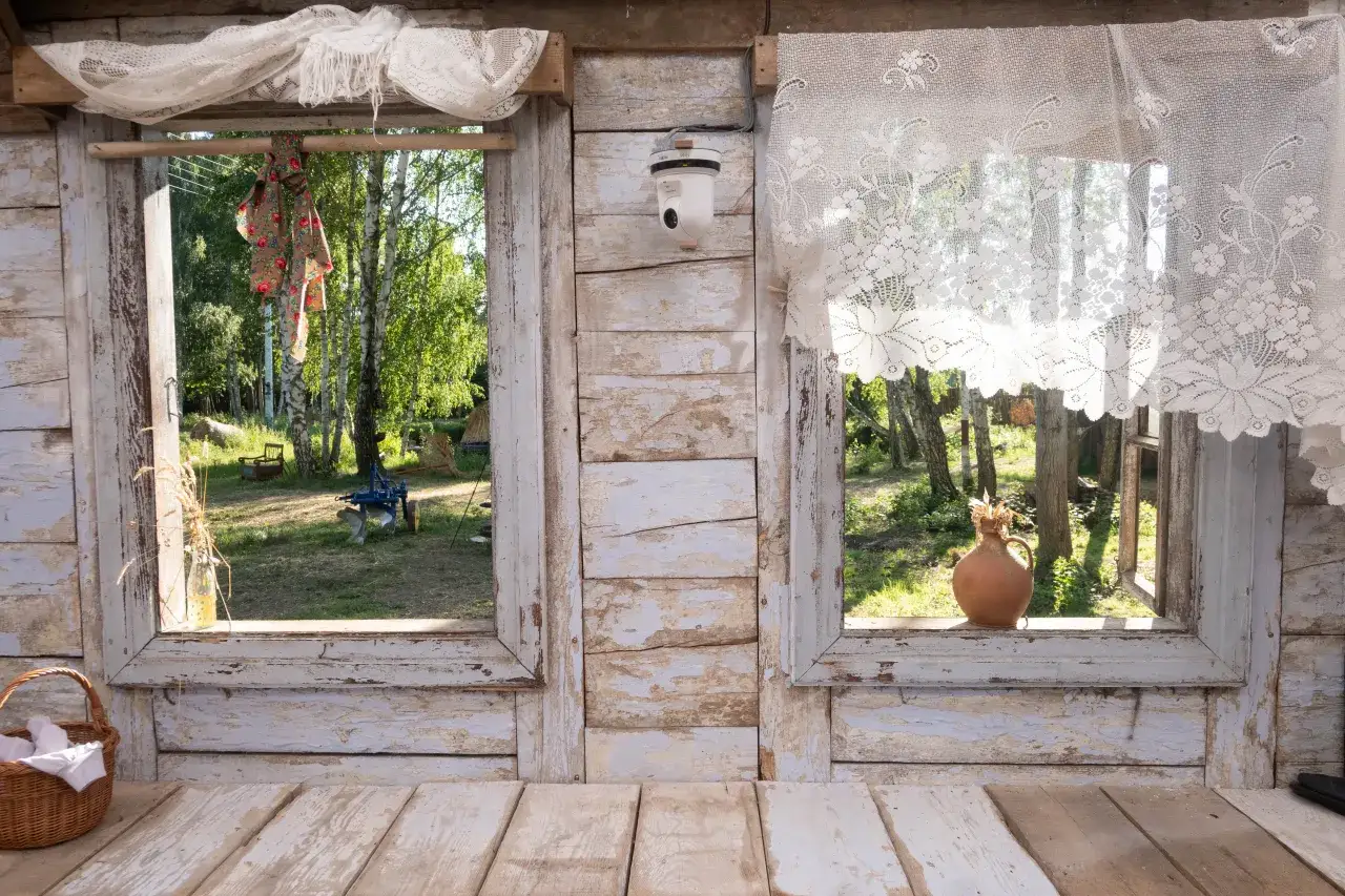 Widok z okna na podwórko, gdzie widać stare narzędzia rolnicze i drzewa. W koszyku leżą białe tkaniny, a na parapecie gliniana waza. To miejsce, gdzie można poczuć zysk z kurnika.