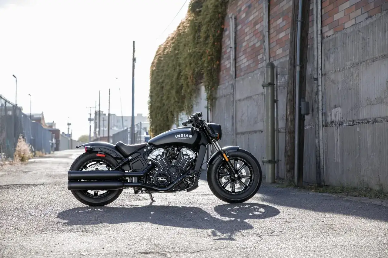 Czarny motocykl cruiser Indian Scout stoi na ulicy, otoczony miejską scenerią z ceglanym murem i zielenią.