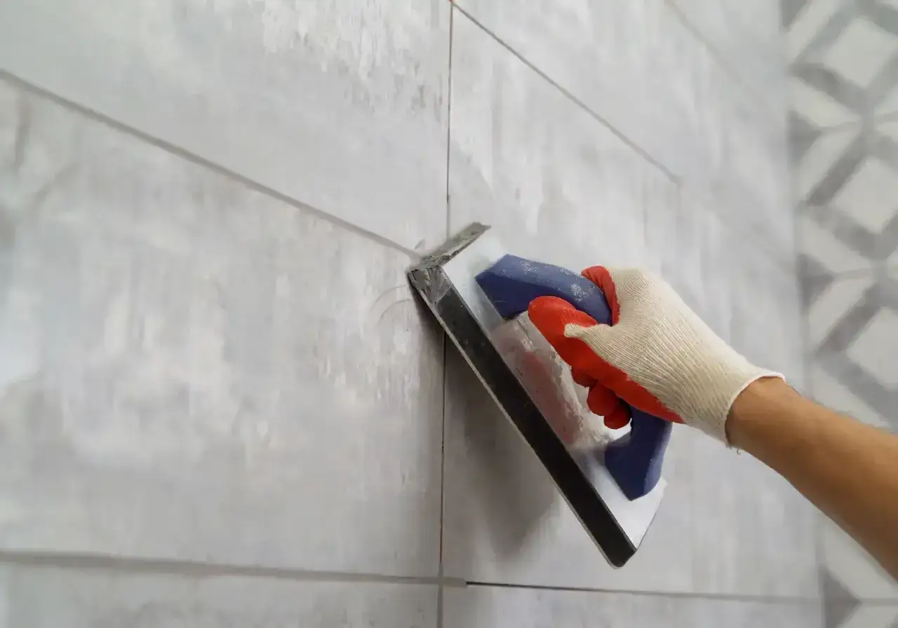 Ręka w rękawicy nakłada fugę na szare płytki za pomocą kielni.