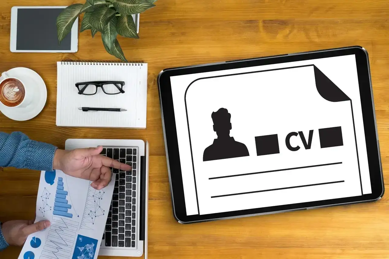 Tablet z ikoną CV i sylwetką mężczyzny, obok laptop, notatnik, okulary i kawa.