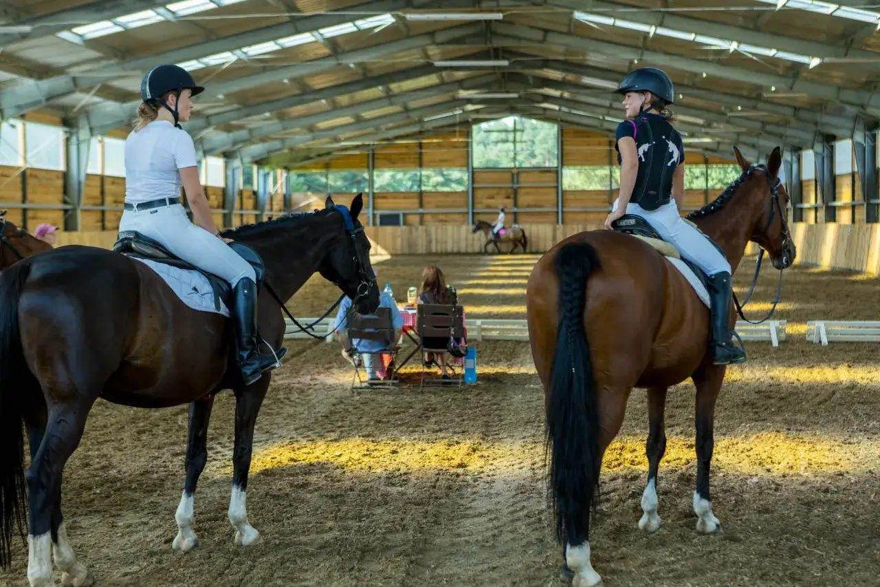 Dwie młode amazonki w kaskach i białych bryczesach siedzą na koniach w krytej ujeżdżalni.
