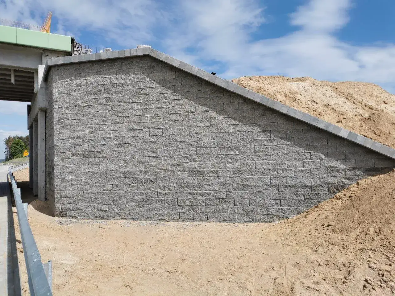 Mur oporowy z szarej kostki brukowej, obok nasypu ziemnego i fragmentu wiaduktu.