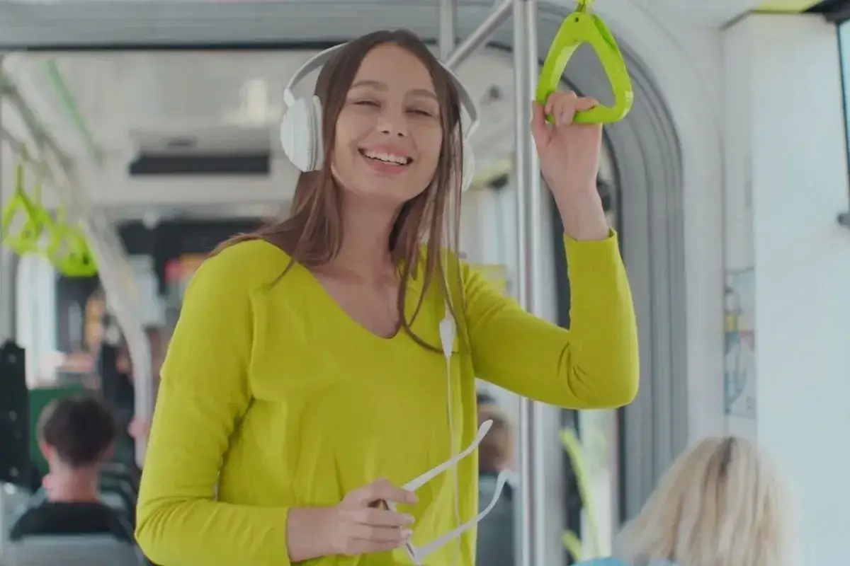 Uśmiechnięta dziewczyna w słuchawkach słucha podcastów po angielsku w tramwaju, trzymając się poręczy.