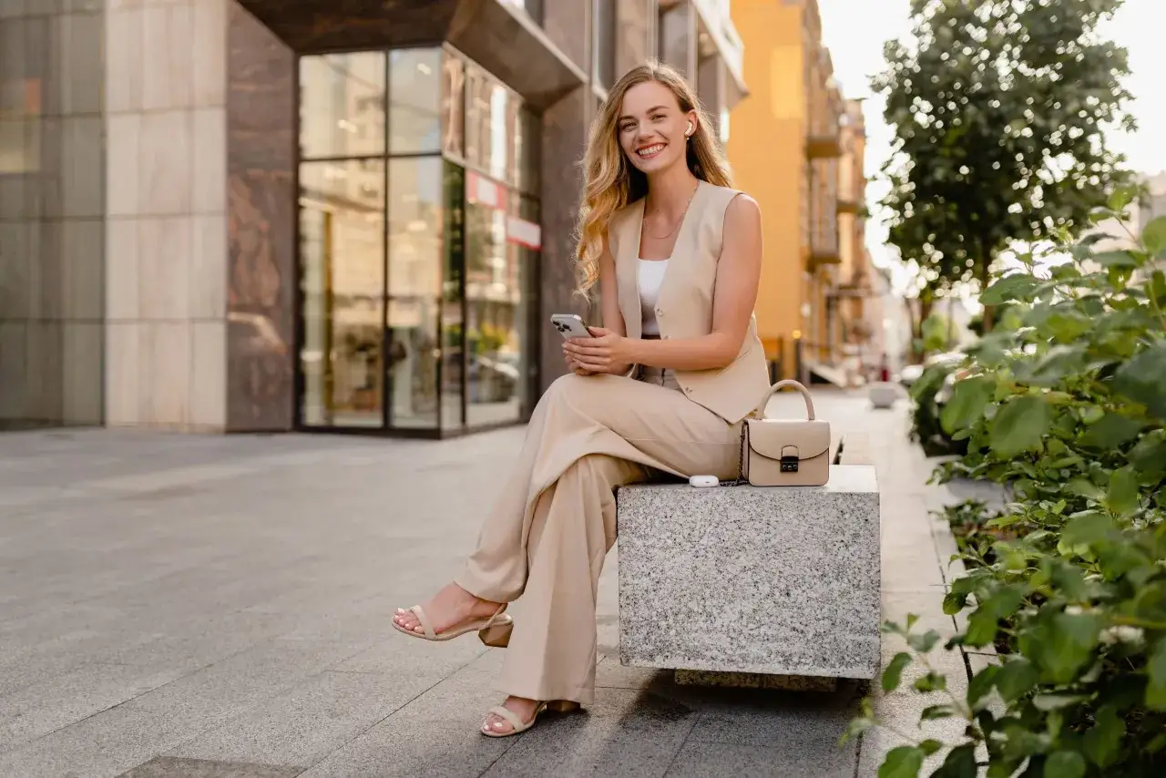 Uśmiechnięta kobieta w beżowym garniturze, idealnym do stylu smart casual, siedzi na ławce z telefonem w dłoni.