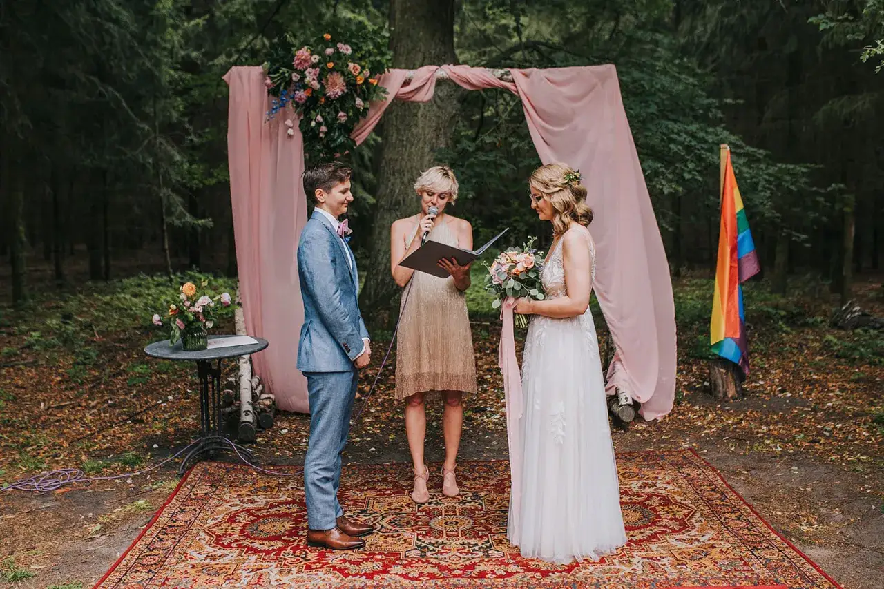 Ceremonia ślubna w lesie, gdzie można wziąć ślub homoseksualny. Dwie kobiety, jedna w garniturze, druga w sukni ślubnej, stoją przed urzędniczką. Tęczowa flaga symbolizuje miłość.
