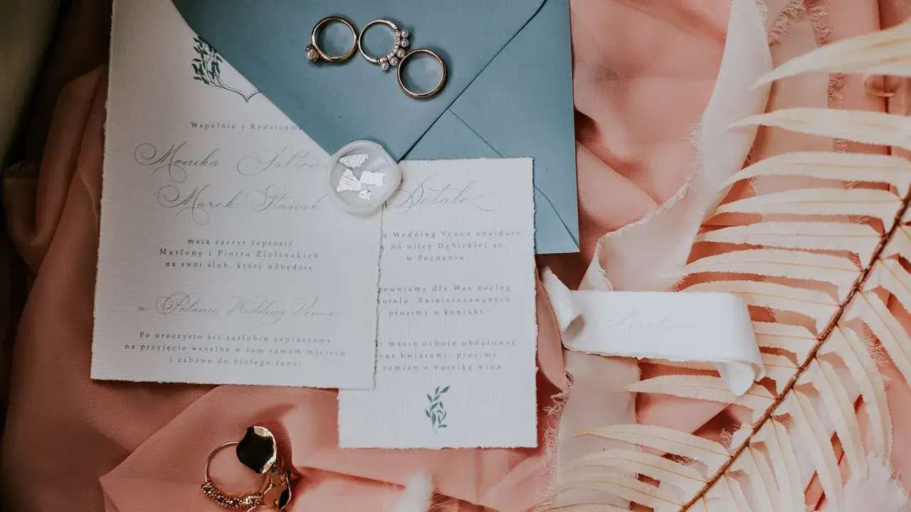 Eleganckie zaproszenia ślubne, obrączki i ozdobne liście na różowym tle.