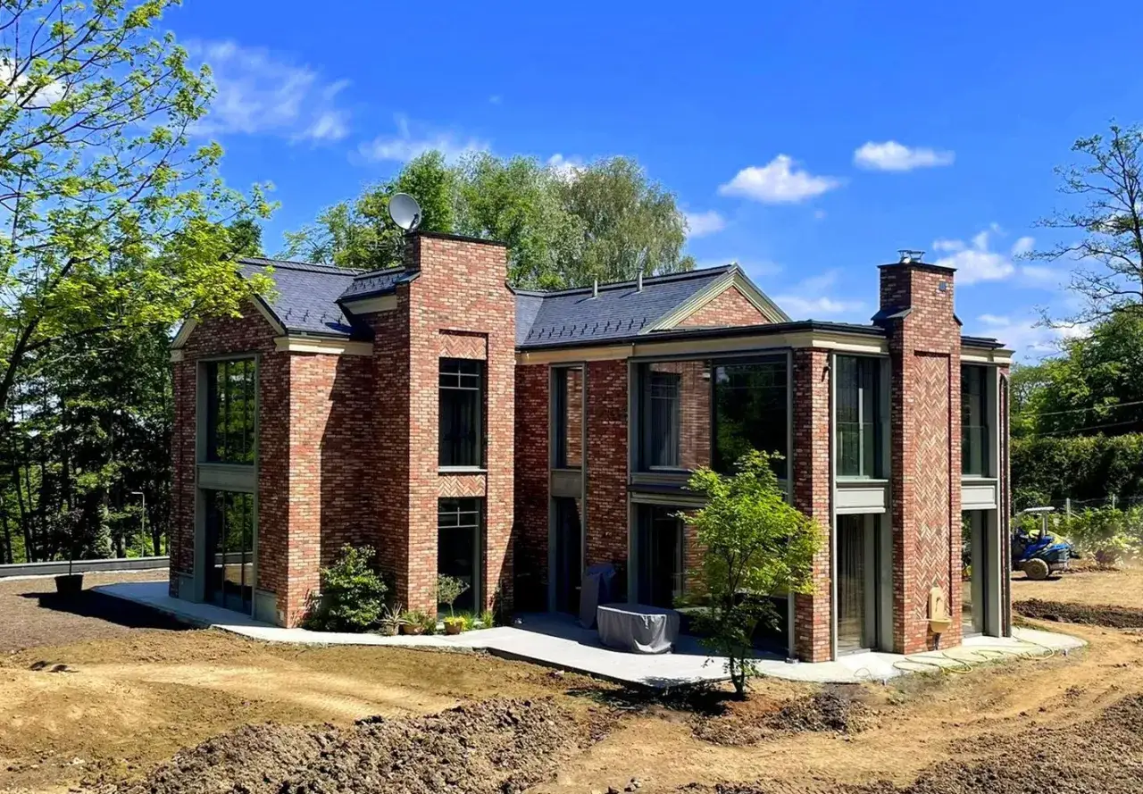 Nowoczesny dom w stylu holenderskim z cegły, z dużymi oknami i zielenią wokół.