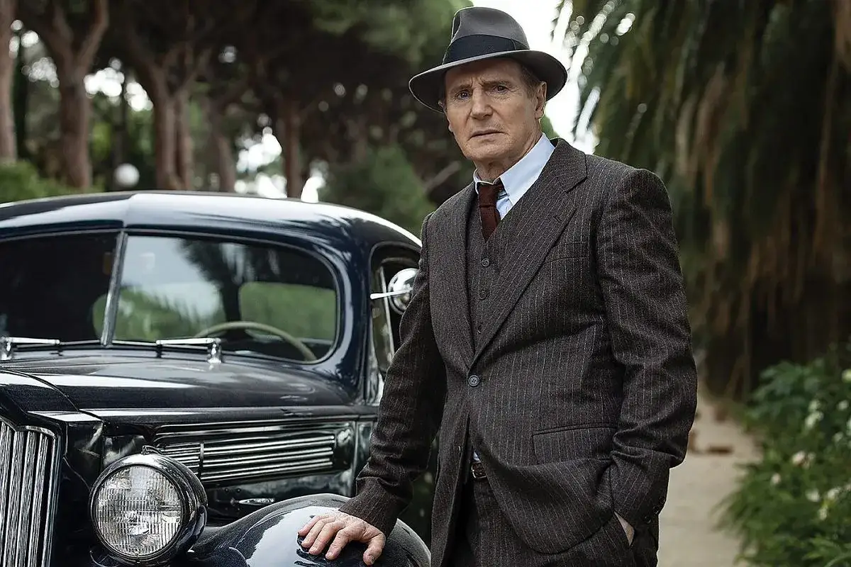 Philip Marlowe, detektyw z kryminałów Raymonda Chandlera, w eleganckim garniturze i kapeluszu, opiera się o luksusowy samochód z lat 40.