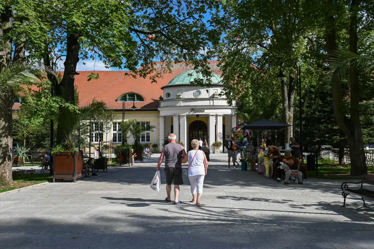 Para starszych osób spaceruje alejką przed budynkiem sanatorium.