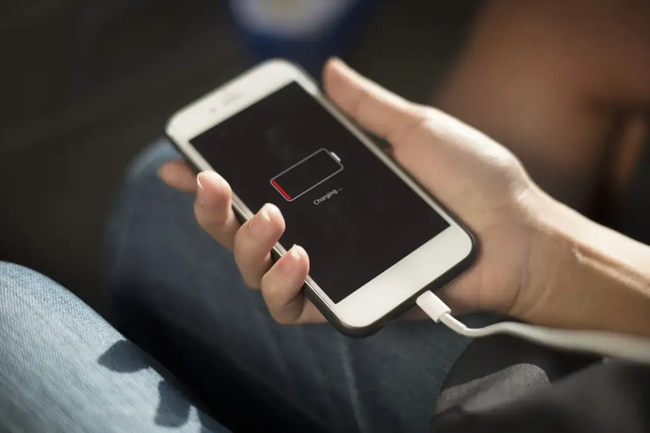 Dłoń trzyma smartfon z ikoną niskiego poziomu baterii i napisem "Charging". Wtyczka kabla ładującego jest podłączona. Oszczędzanie baterii jest kluczowe.