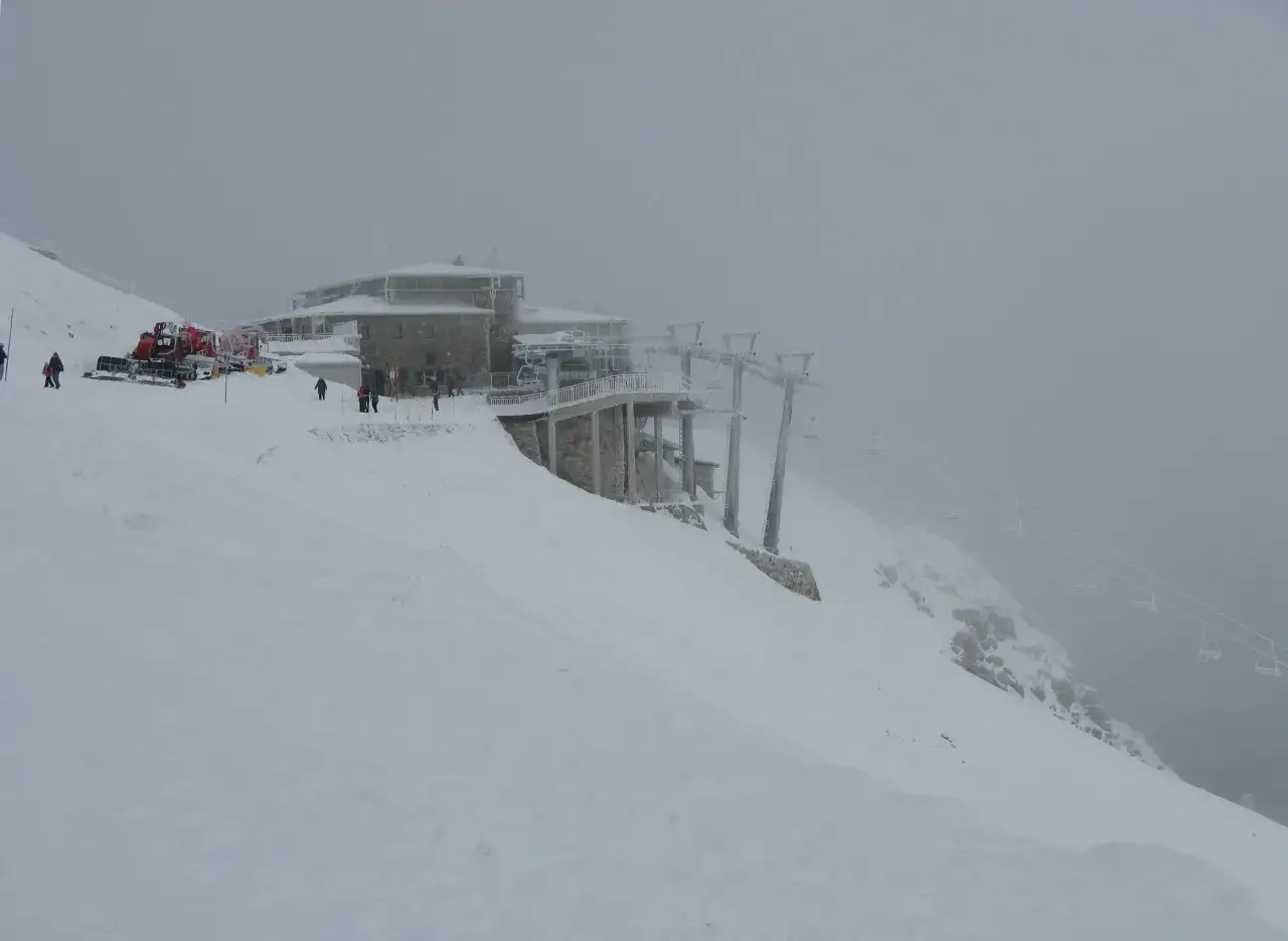 Mglisty dzień na Kasprowym Wierchu. Kolejka krzesełkowa wjeżdża na szczyt, a ratrak przygotowuje trasę do narciarstwa.
