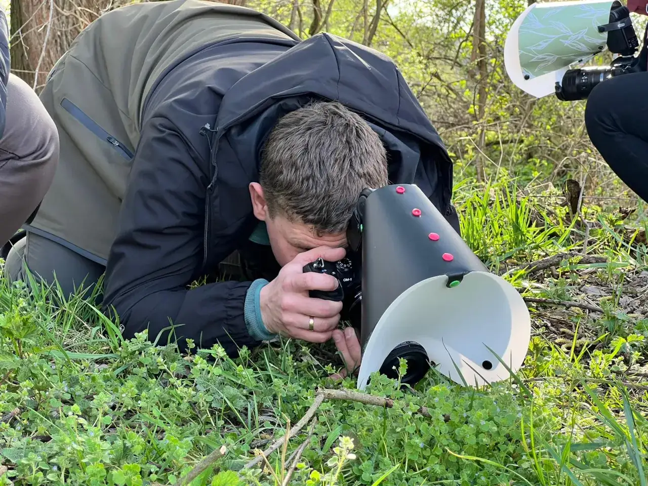 Fotograf z aparatem i dyfuzorem do makro pochyla się nad trawą, by uchwycić drobne detale natury.