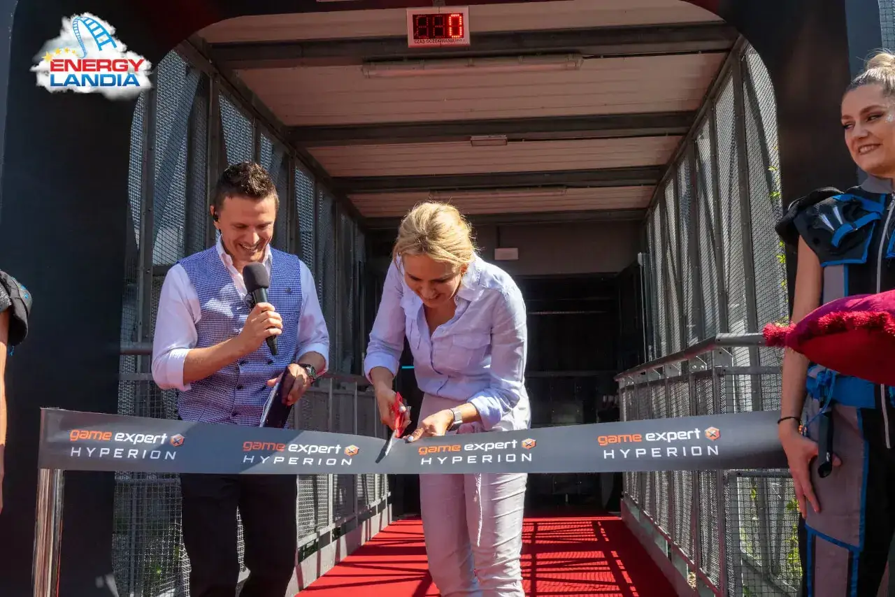 Kobieta przecina wstęgę z napisem "game expert HYPERION", mężczyzna trzyma mikrofon.