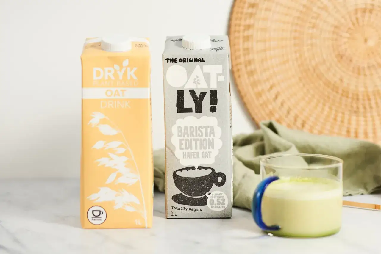 Dwa kartony mleka roślinnego (DRYK i Oatly) oraz szklanka zielonego napoju.
