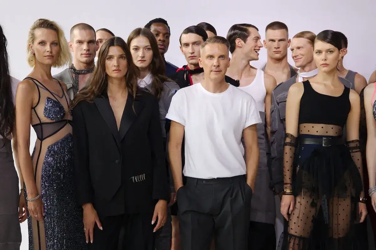 Grupa modeli i modelek pozuje obok słynnych projektantów mody. W centrum mężczyzna w białej koszulce, obok kobiety w czarnej marynarce.