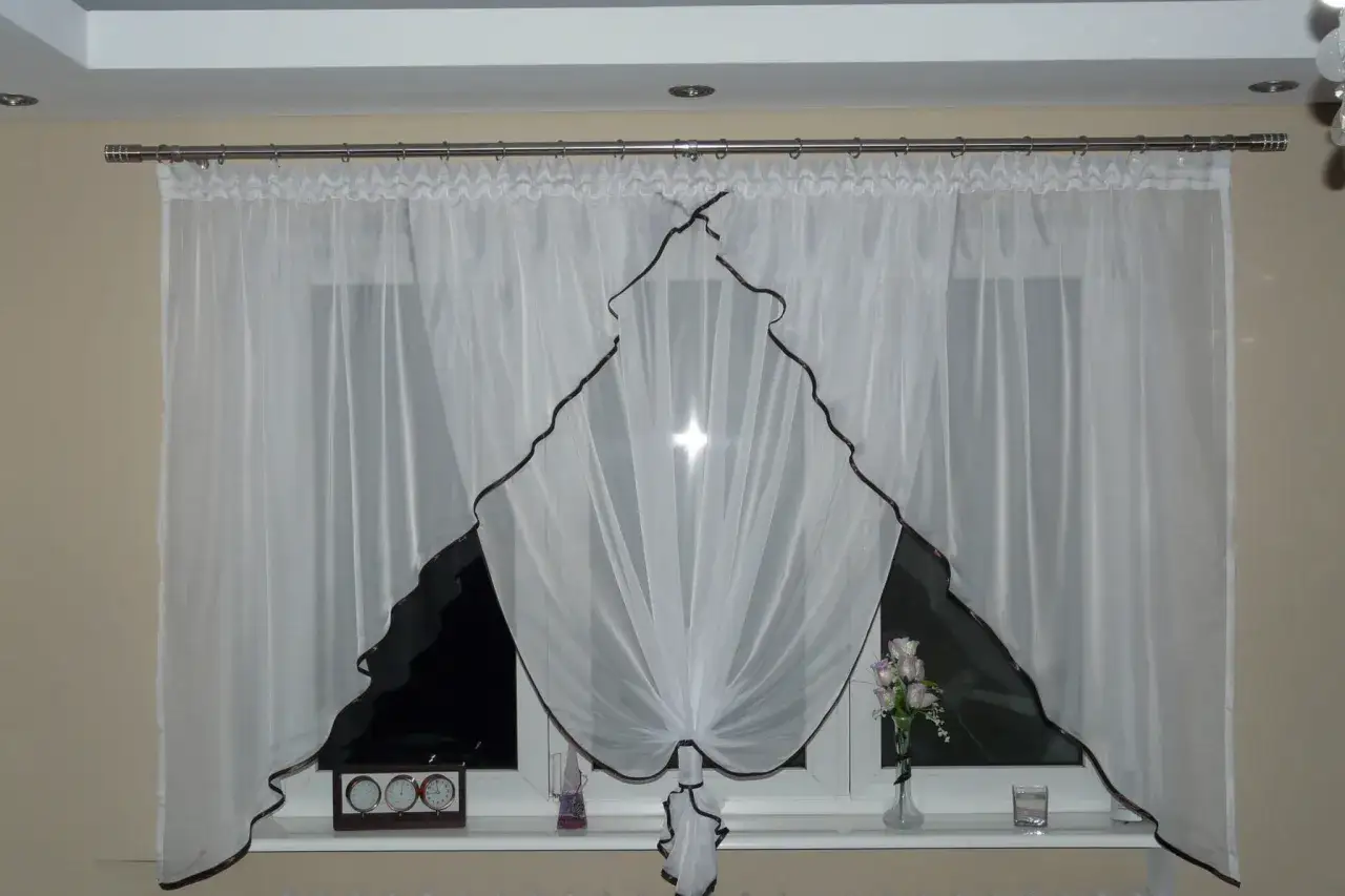 Eleganckie białe firanki z brązową lamówką, idealnie pasujące na karnisz 2m. Na parapecie zegar i wazon z kwiatami.