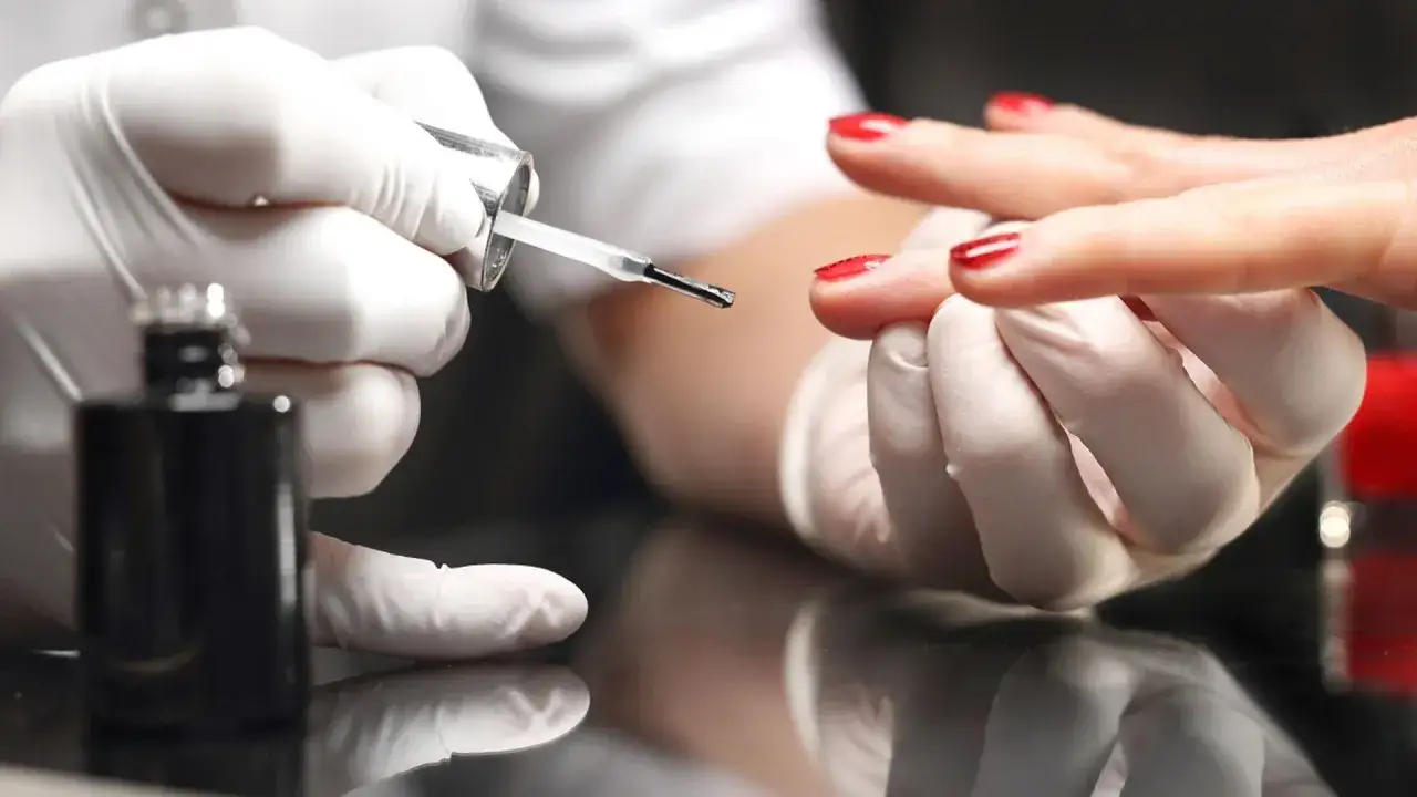 Dłoń w białej rękawiczce aplikuje czerwony lakier. Uważaj na uczulenie na hybrydę, wybieraj bezpieczne produkty.