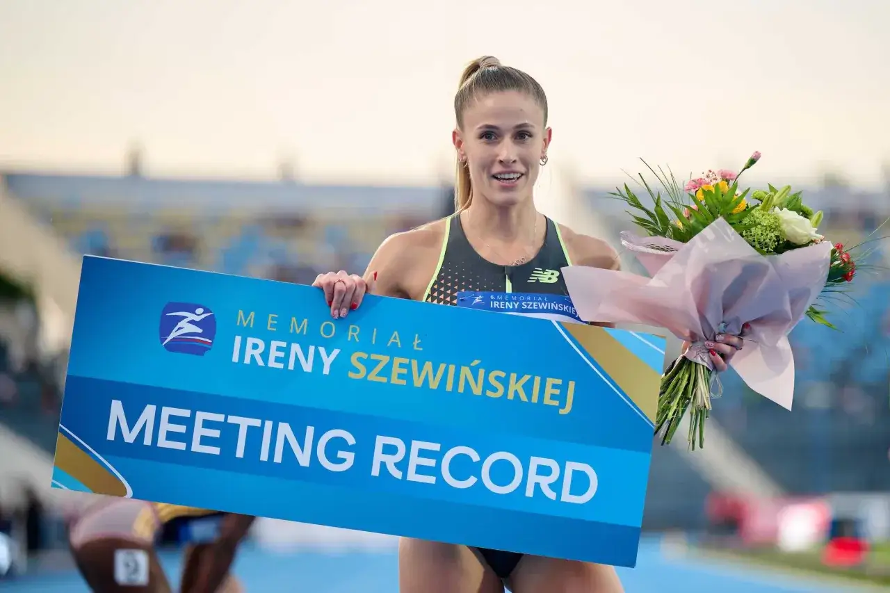 Uśmiechnięta lekkoatletka z bukietem kwiatów trzyma tablicę "MEETING RECORD" z logo memoriału Ireny Szewińskiej.