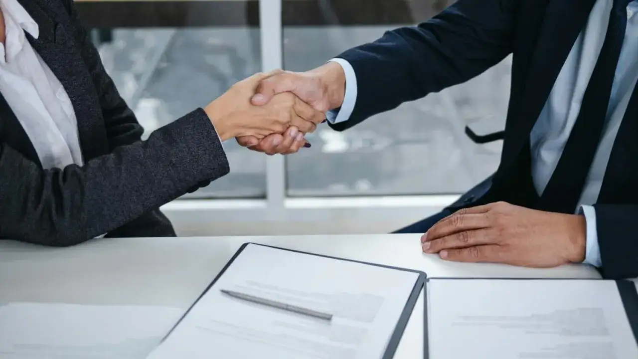 Dwie osoby w garniturach ściskają sobie dłonie nad stołem z dokumentami.