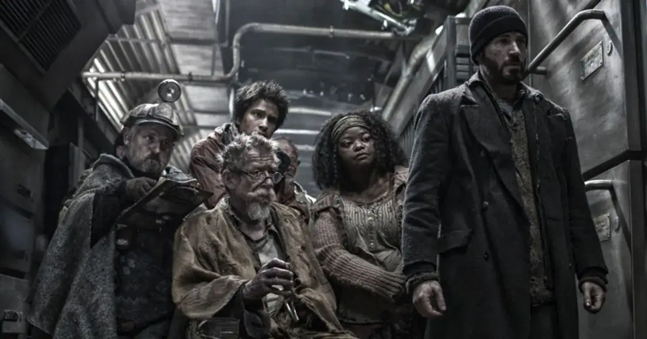 Grupa pasażerów Snowpiercer: Arka Przyszłości, w tym mężczyzna w czapce i płaszczu, siedzi w ciasnym wagonie.