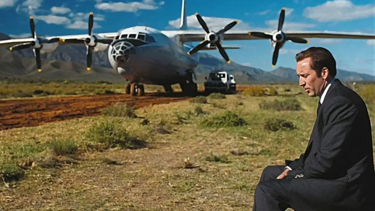 Mężczyzna w garniturze siedzi na tle samolotu transportowego. W jego oczach widać ciężar, jakby był panem życia i śmierci.