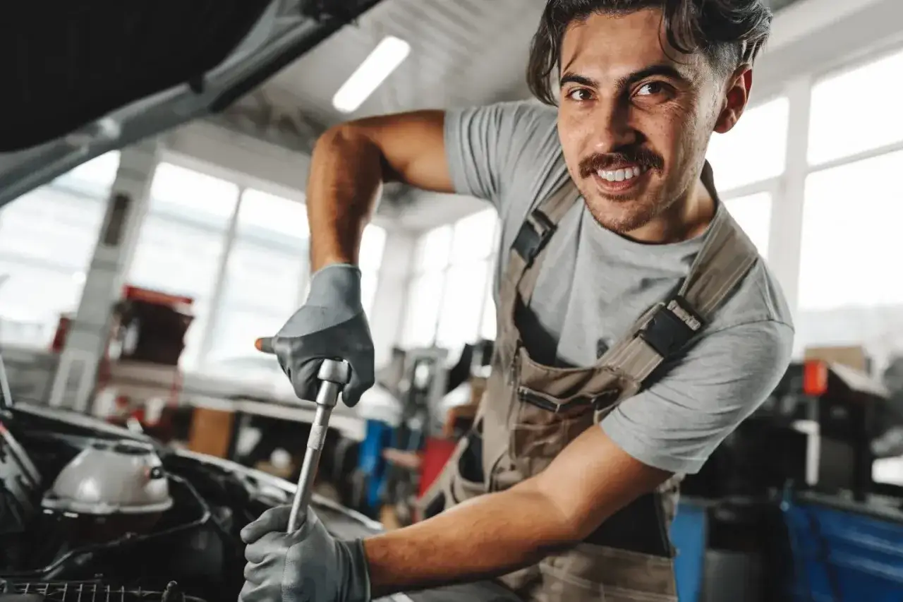 Mechanik w ogrodniczkach i rękawiczkach naprawia silnik samochodu kluczem nasadowym.