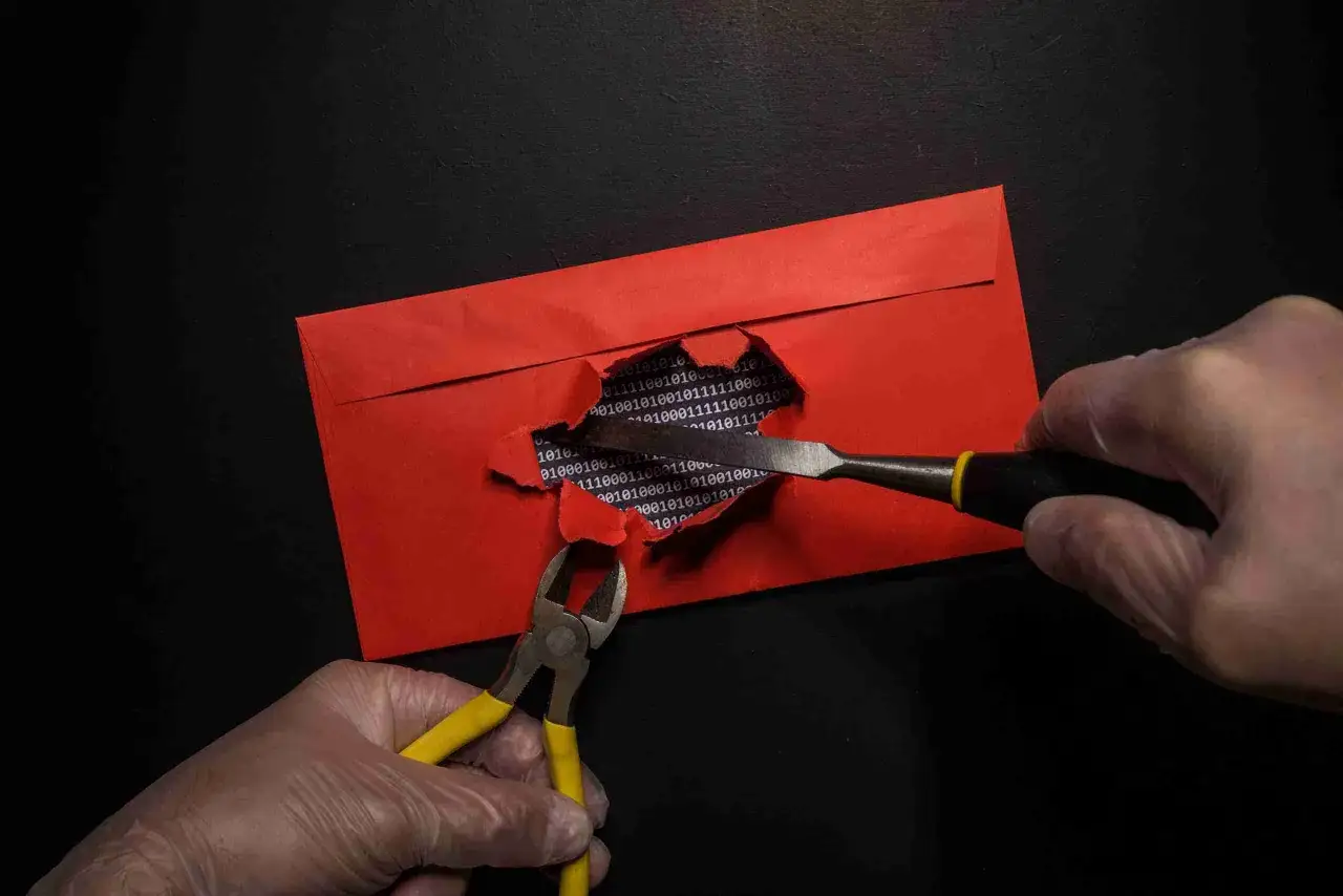 Ręce w rękawiczkach rozdzierają czerwianą kopertę, odsłaniając kod binarny. Narzędzia sugerują wektor ataku na dane.