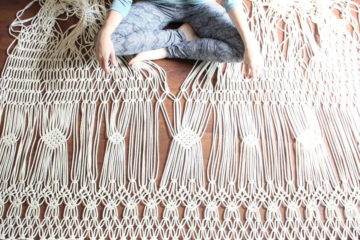Osoba siedzi na podłodze, tworząc skomplikowane makrama wzory. Widać misternie splecione sznurki, tworzące geometryczne kształty.