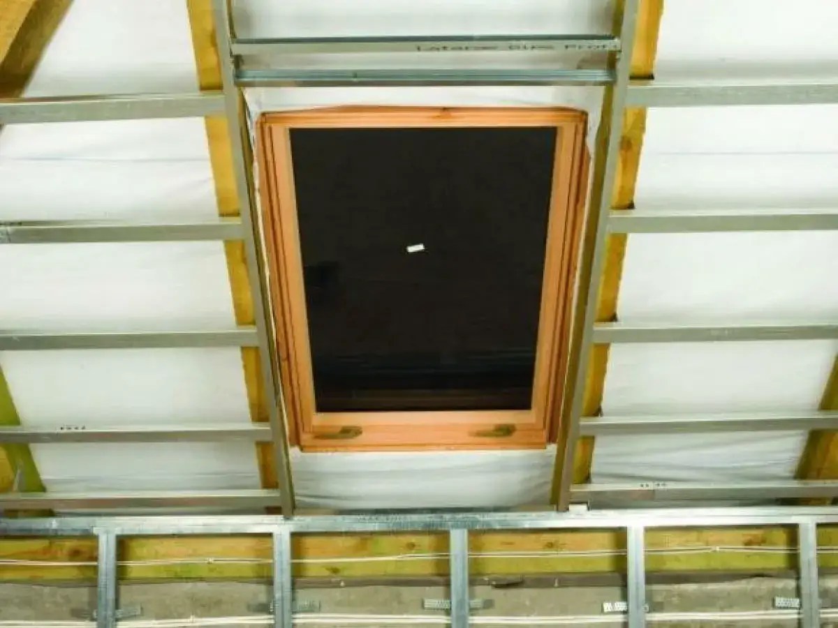 Ocieplanie okna dachowego karton gipsem. Widok na drewniane okno dachowe w trakcie prac wykończeniowych poddasza, z widoczną konstrukcją metalową i izolacją.