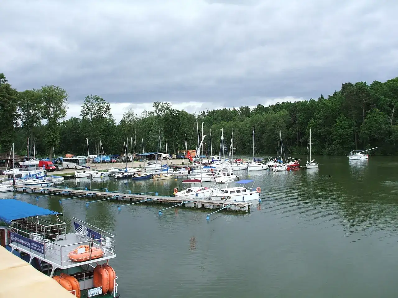 Port w Rucianem Nidzie pełen żaglówek i łodzi. W tle las i pochmurne niebo.